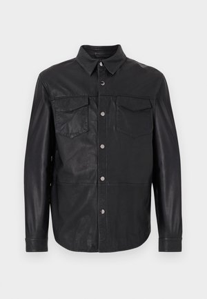 Mann trägt schwarze, geknöpfte Lederjacke, passende Lederhose und Sonnenbrille, steht vor hellem einfarbigem Hintergrund.