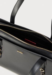 Sac à main en cuir noir avec ferrures dorées, double poignée et fermeture éclair sur le dessus. Intérieur doté d'une étiquette logo rouge et d'une doublure lisse.