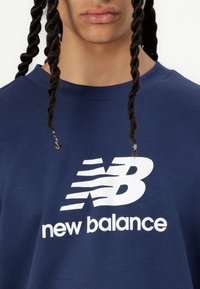 Navy zila kokvilnas džemperis ar apaļu apkakli, ar lielu baltu "New Balance" logotipu un svītrām virs burtiem.