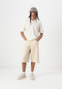 Beige shorts, teksturert hvit kortermet skjorte og lyse joggesko. Modell iført en grå caps med logo, stående mot en hvit bakgrunn.