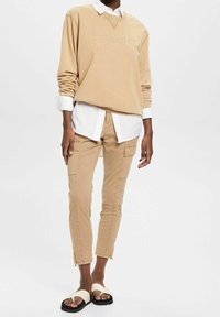 Beige bomullströja med prägling av logotyp, lager på vit kragskjorta. Bruna cargobyxor med sidofickor, ihop med krämfärgade sandaler.