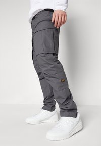 Šedé cargo kalhoty s několika kapsami a zipovým designem, vyrobené z odolné látky. Nosené s bílými sportovními teniskami.
