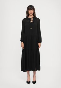 Abito maxi nero realizzato in tessuto leggero. Presenta un collo a volant, maniche lunghe e una gonna a strati. Abbinato a tacchi neri a punta.