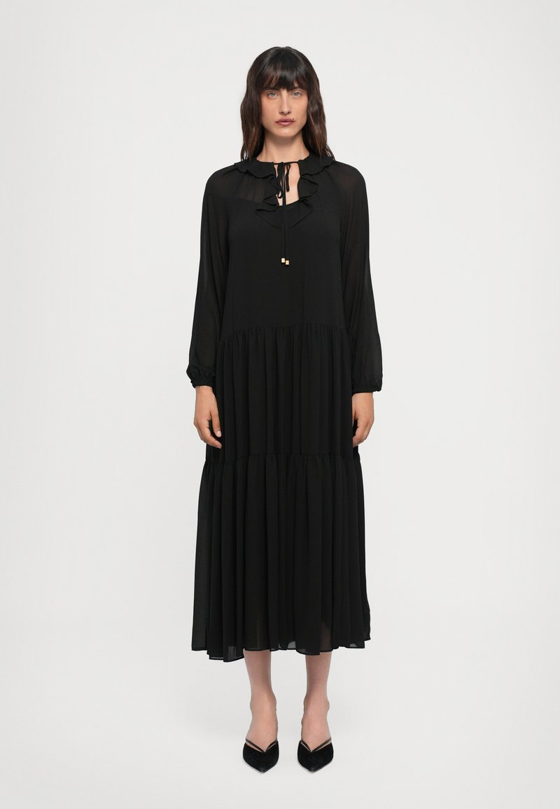 Abito maxi nero realizzato in tessuto leggero. Presenta un collo a volant, maniche lunghe e una gonna a strati. Abbinato a tacchi neri a punta.