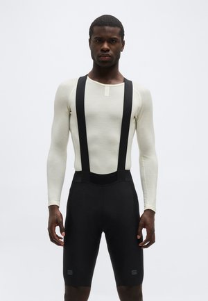 Egy férfi, aki testhez simuló hosszú ujjú fehér inget és fekete kerékpáros rövidnadrágot visel, amely tartópántokkal van ellátva, egy egyszerű háttér előtt áll.