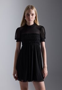 Abito nero con maniche a sbuffo trasparenti e corpetto arricciato. Gonna fluida con una consistenza morbida, caratterizzata da un collo alto e un taglio al ginocchio.