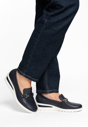 Füße in marineblauen Leder-Slips mit goldener Schnallenverzierung und weißen Gummisohlen, kombiniert mit dunklen Jeans.