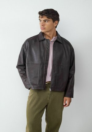 Dunkelbraune Lederjacke mit einem Kragen, zwei vorderen Taschen und einem Reißverschluss. Getragen über einem hellrosa Oberteil und olivgrünen Hosen.