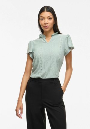 Femme debout portant un chemisier vert clair à manches courtes avec un col volanté et un pantalon noir taille haute, les mains dans les poches.