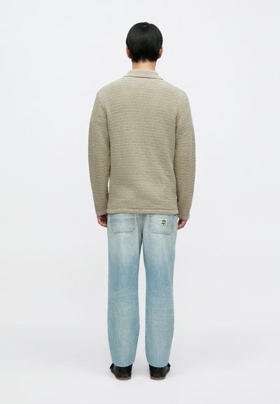Béžový pletený sveter s textúrovaným vzorom, s golierovým výstrihom, v kombinácii so svetlomodrými džínsami s rovnými nohavicami a čiernymi topánkami.