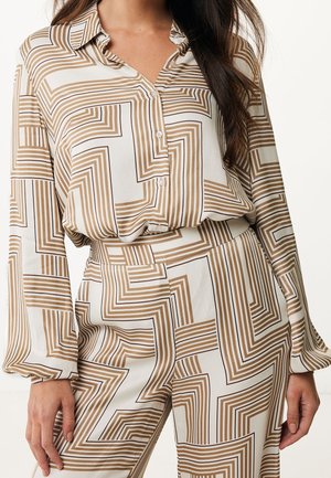 Vrouw draagt een witte blouse en broekset met bruine geometrische doolhofachtige strepen, toont torso en armen, geen zichtbaar hoofd.