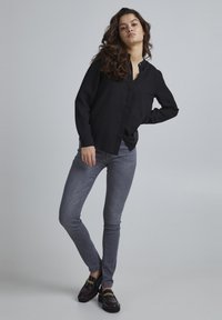 Zwarte blouse met lange mouwen en een knoopsluiting aan de voorkant, gecombineerd met lichtgrijze slim fit jeans en donkere instappers met een gouden kettingaccent.