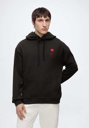 Giovane uomo con felpa nera con cappuccio con ricamo di rosa rossa sul petto e pantaloni bianchi, in piedi con una mano sui fianchi contro uno sfondo semplice.