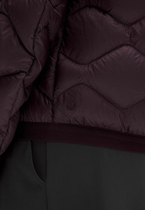 Ciemnofioletowa pikowana kurtka z założonym dołem na czarne spodnie z boczną kieszenią, pokazująca subtelne logo marki na materiale kurtki.