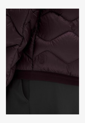 Jachetă matlasată de culoare purpurie închisă, cu tivul peste pantaloni negri cu buzunar lateral, având un logo subtil al brandului pe țesătura jachetei.