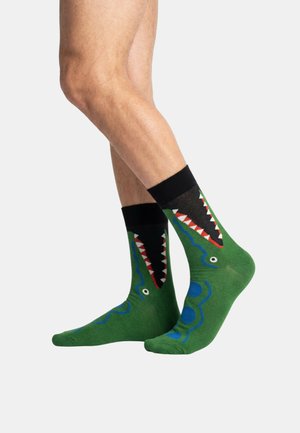 Beine mit grünen Socken mit schwarzen Bündchen und einem Design eines offenen Krokodilmundes mit weißen Zähnen auf einfarbigem Hintergrund.