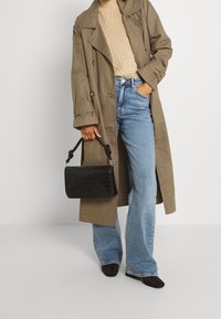 Person, der einen beigen Trenchcoat, einen cremefarbenen Strickpullover, weite blaue Jeans, schwarze Schuhe trägt und eine schwarze Handtasche hält.