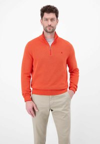 Oranges Strickstrickjacke mit Reißverschluss und strukturiertem Muster, ausgestattet mit hohem Kragen und gerippten Bündchen. Kombiniert mit beigen Hosen.