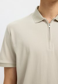 Homme portant un polo beige à manches courtes avec col et bords de manches texturés, fermeture éclair frontale partiellement ouverte, raccourci au niveau du cou et de l'épaule.
