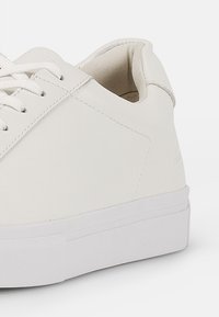 Valkoiset sneakerit, joissa on sileä nahkapinta, suorat valkoiset nauhat ja paksu valkoinen kumipohja, jossa on minimalistiisia muotoiluelementtejä.