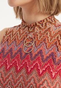 Ärmelloses Oberteil mit hohem Kragen, das ein Zickzackmuster in Orangetönen, Rot und Blau zeigt, mit einem metallischen Ringdetail am Kragen.