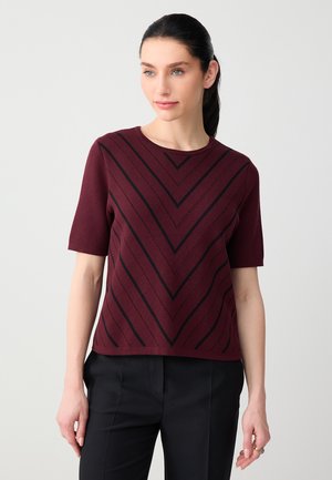 Kobieta o ciemnych włosach związanych w kucyk, ubrana w bordowy sweter z krótkim rękawem z czarnymi pasami w kształcie litery V oraz czarne spodnie, stojąca na jasnym, jednolitym tle.