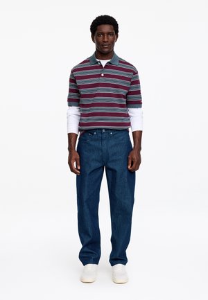 Mężczyzna w bordowo-szarej koszulce polo w paski na białej bluzce z długim rękawem, niebieskich dżinsach i białych trampkach, stojący na jednolitym tle.