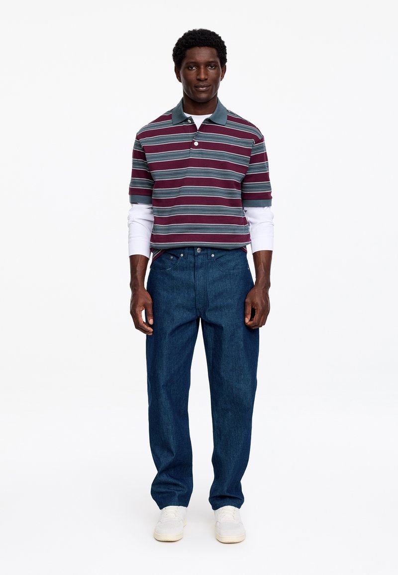 Homme portant un polo rayé bordeaux et gris sur un t-shirt à manches longues blanc, un jean bleu et des baskets blanches, debout devant un fond uni.