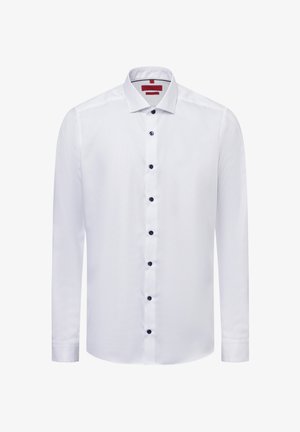 Weißes langärmliges Hemd aus strukturiertem Stoff, mit klassischem Kragen und dunkelblauen Knöpfen, die vorne entlang laufen. Rotes Etikett im Kragen.
