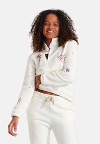 Pullover in pile bianco sporco con apertura a zip sulla metà, ricami floreali e polsini a coste. Abbinato a pantaloni coordinati con coulisse.