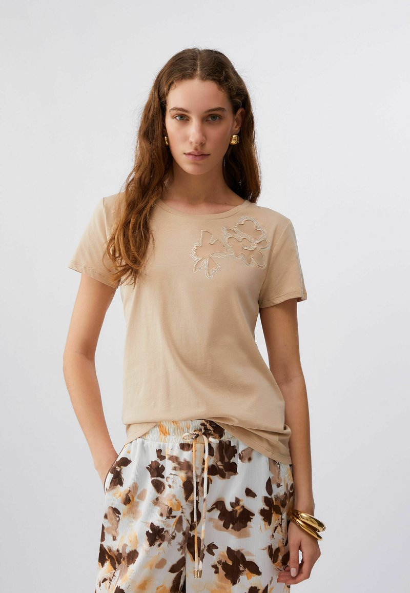 Camiseta de algodón beige con mangas cortas, con diseño floral recortado y detalles con cuentas en el lado izquierdo; textura suave.