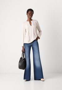 Beige bluse med nøkkelhullsdetalj og folder, kombinert med vide blå denimjeans. Modellen holder en svart håndveske og har på seg lyse sko.