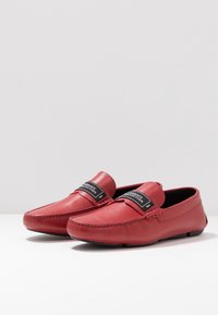 Rote Lederloafers verfügen über gestickte Details und ein breites schwarzes Markenlabel auf der Vorderseite. Die Sohle ist flach und flexibel.