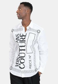 Witte blouse met knoopjes, voorzien van zwarte grafische prints, waaronder een touwontwerp en tekst. Gemaakt van gladde stof met een klassieke kraag.