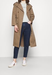 Beżowy trencz z naramiennikami, zapięciem na guziki i luźnym krojem, noszony na jasnej zielonej koszuli i ciemnoniebieskich dżinsach z białymi sneakersami.