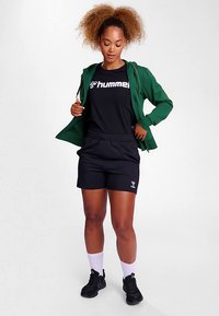 Groene jas, zwart T-shirt met logo, zwarte korte broek, witte sokken en zwarte sportschoenen. Model staat met handen in de zakken.