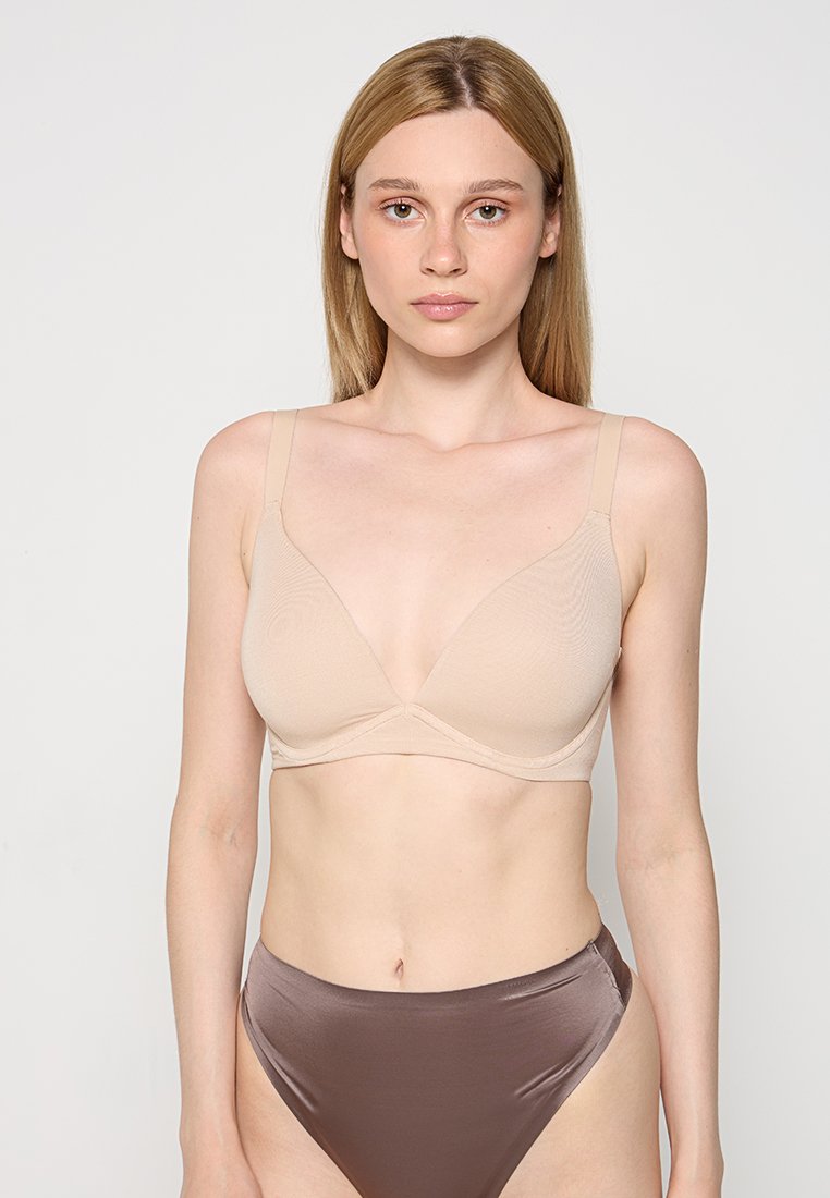 Beżowy, bezprzewodowy biustonosz z gładkim, trójkątnym krojem miseczek oraz regulowanymi ramiączkami. Miękka tkanina zapewnia subtelną teksturę i komfortowe dopasowanie.