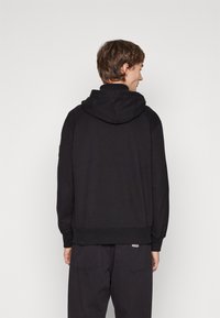 Sudadera negra con cordón y corte relajado. Hecha de tela suave, cuenta con puños y dobladillo acanalados. La vista trasera muestra un diseño sin costuras.