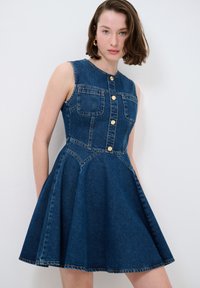 Robe sans manches en denim bleu foncé, avec un corsage ajusté comprenant deux poches avant, des boutons dorés et une jupe évasée.