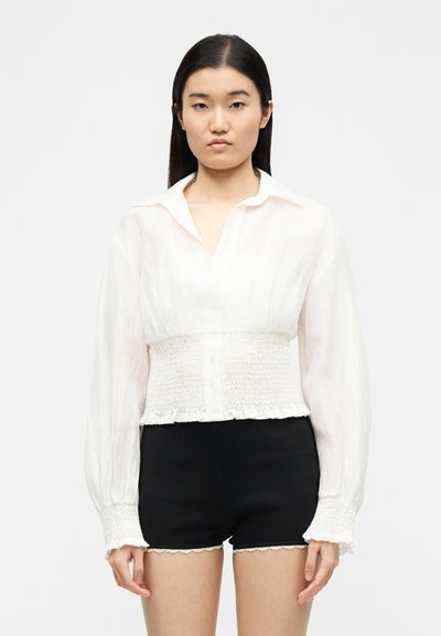 Jeune femme aux cheveux noirs raides portant un chemisier blanc à manches longues smocké et un short noir avec un galon en dentelle, debout devant un fond uni.