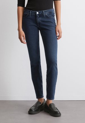 Jeans en denim bleu foncé avec une coupe slim, présentant un design classique à cinq poches, un léger délavage et une taille mi-haute. Portés avec des mocassins noirs.