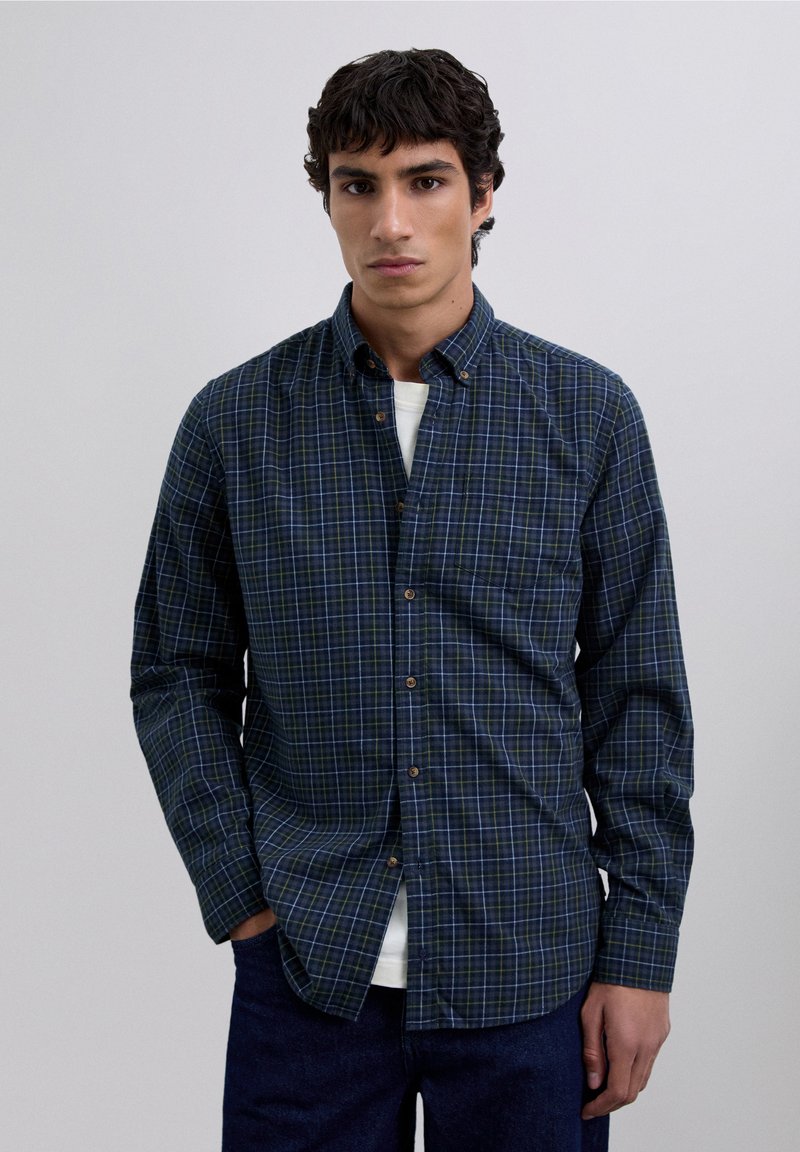 Camisa de cuadros azul marino con cuello abotonado, con acentos en verde y blanco, mangas largas y un corte relajado. Botones marrones adornan la parte delantera.
