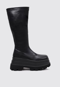 Botas negras hasta la rodilla hechas de cuero suave, con una suela de plataforma gruesa de textura rugosa y costuras visibles en la parte superior.