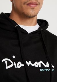 Detailní záběr osoby v černé mikině s kapucí, na které je nápis "Diamond Supply Co." a pod tím bílá košile.