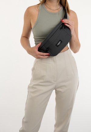 Banane en tissu noir à la forme rectangulaire élégante avec une poche zippée à l'avant, portée en bandoulière sur un haut vert clair.