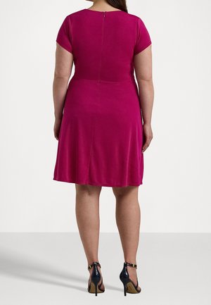 Kvinde stående med ryggen mod, iført en kortærmet magenta kjole med lynlås bagpå og sorte højhælede sko med ankelrem.