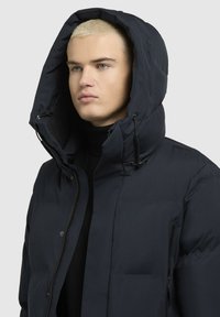 Tengerészkék puffernadrág nagyméretű kapucnival, cipzáros záródással és oldal zsebekkel, sima, időjárásálló anyagból készült.