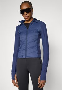 Mėlyna izoliuota striukė su užtrauktuku priekyje ir kapišonu, su V formos raštu ant krūtinės ir rankogaliais su nykščio angomis. Dėvima ant juodų tamprių.