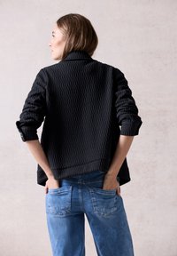 Giacca nera texture con colletto e maniche arrotolate, abbinata a jeans in denim blu chiaro con tasche posteriori.