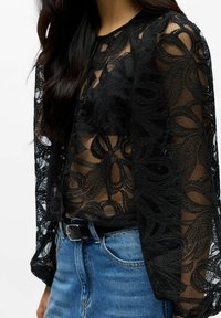 Sort blondebluse med lange, gennemsigtige ærmer, der har indviklede blomsterudskæringer og knapdetaljer, kombineret med et denimskørt og bælte.